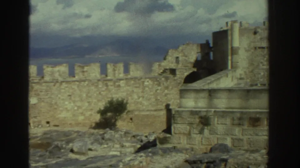 1976 Greece travel footage frame with a stone castle wall, broken tower structures, rocky terrain, and distant water or hills.