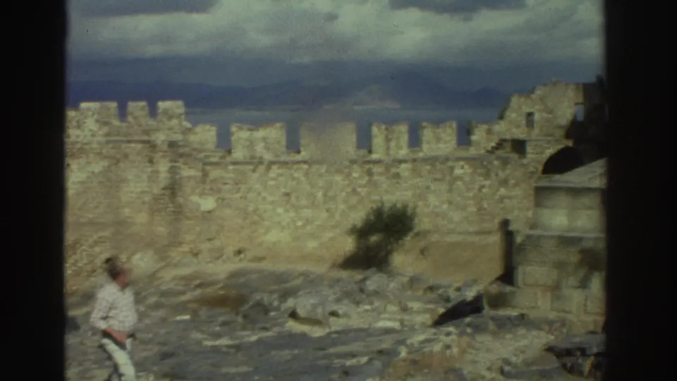 1976 Greece 8mm frame showing an adult tourist in light clothing walking through rocky castle grounds below battlements.