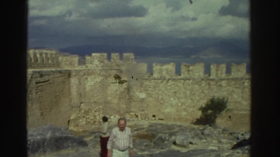 1976 Greece frame showing two adult tourists in castle ruins, with one figure in a red skirt near stone battlements and cloudy hills.