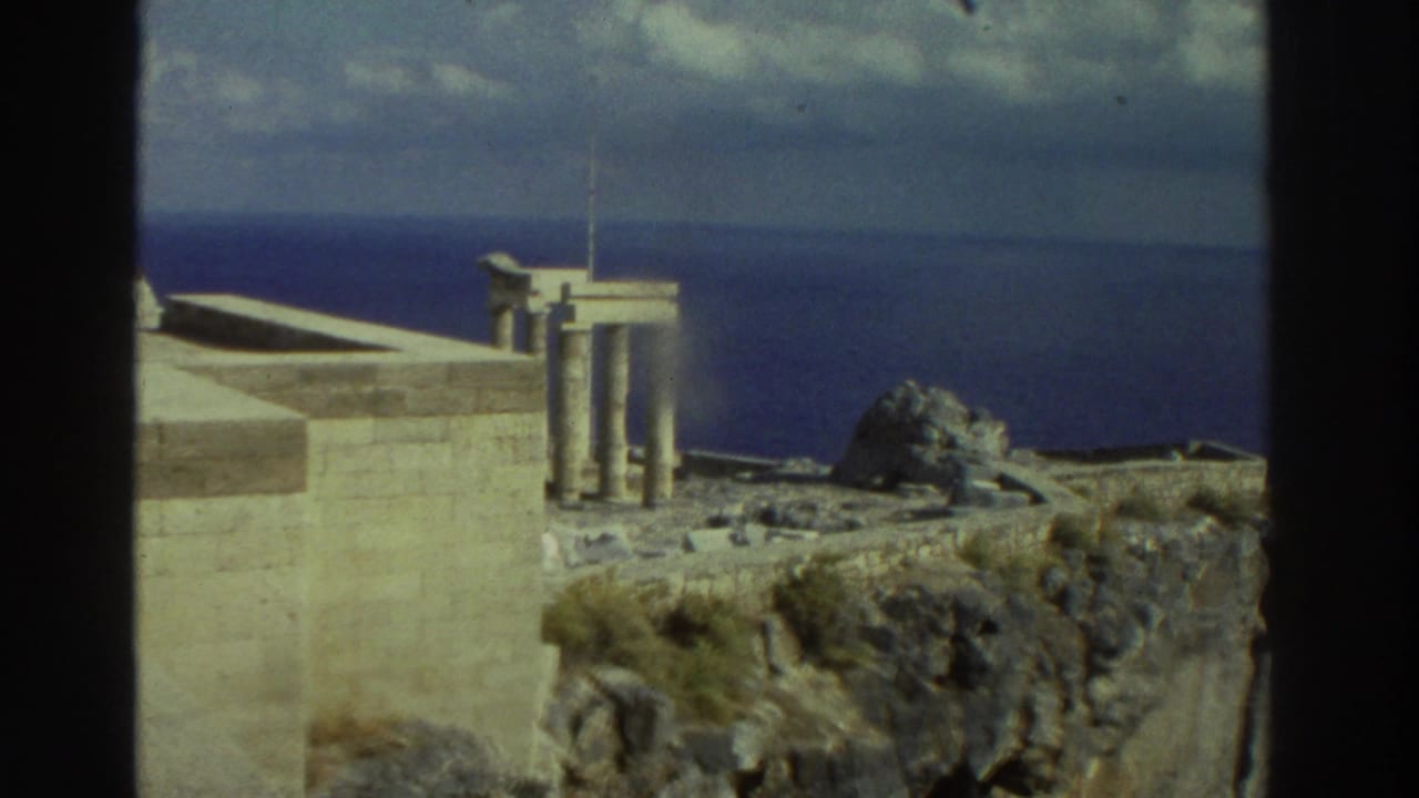 1976 Greece home-movie frame showing pale stone ruins and columns on a cliff edge overlooking deep blue water.