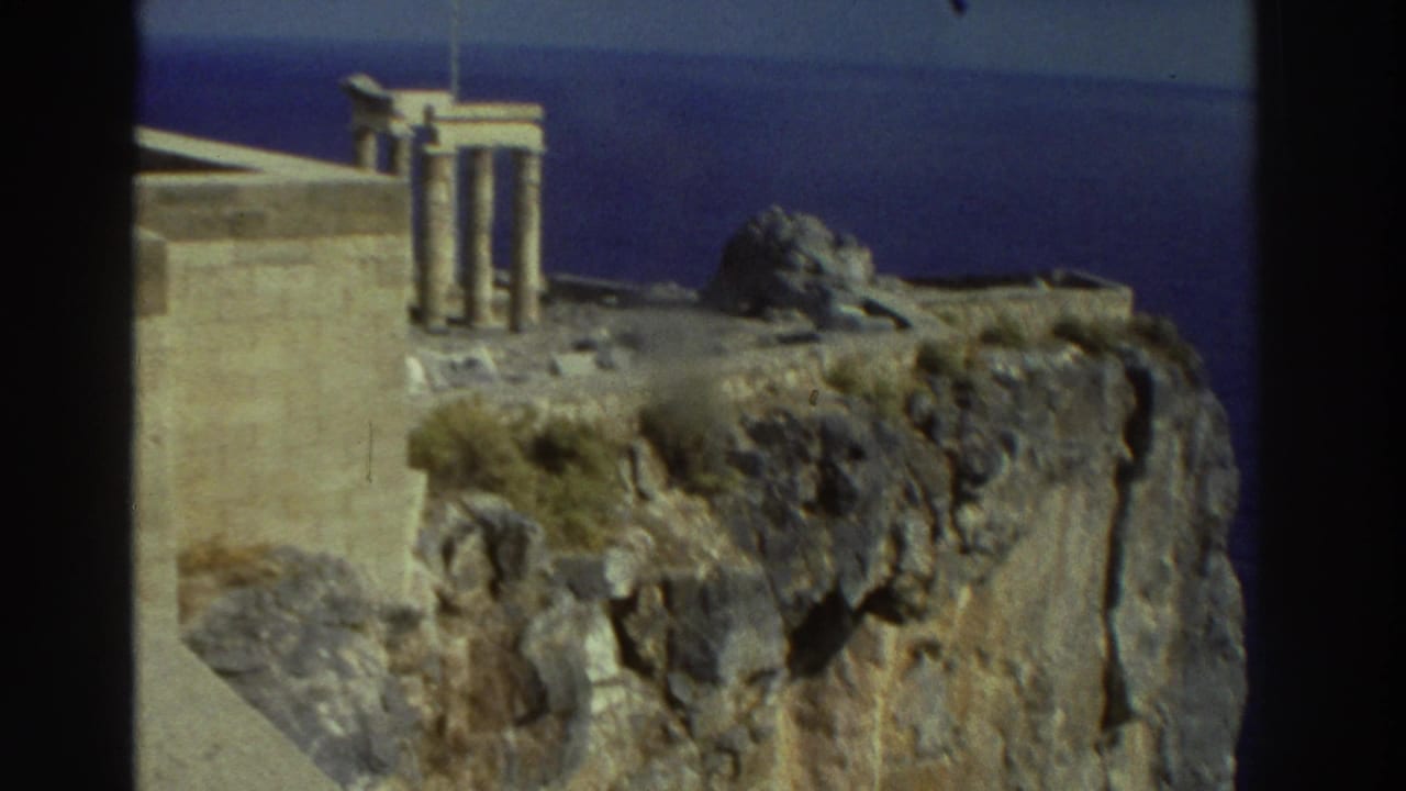 Handheld 8mm view of Greek coastal ruins, with columned stonework above a rugged cliff and sea beyond.