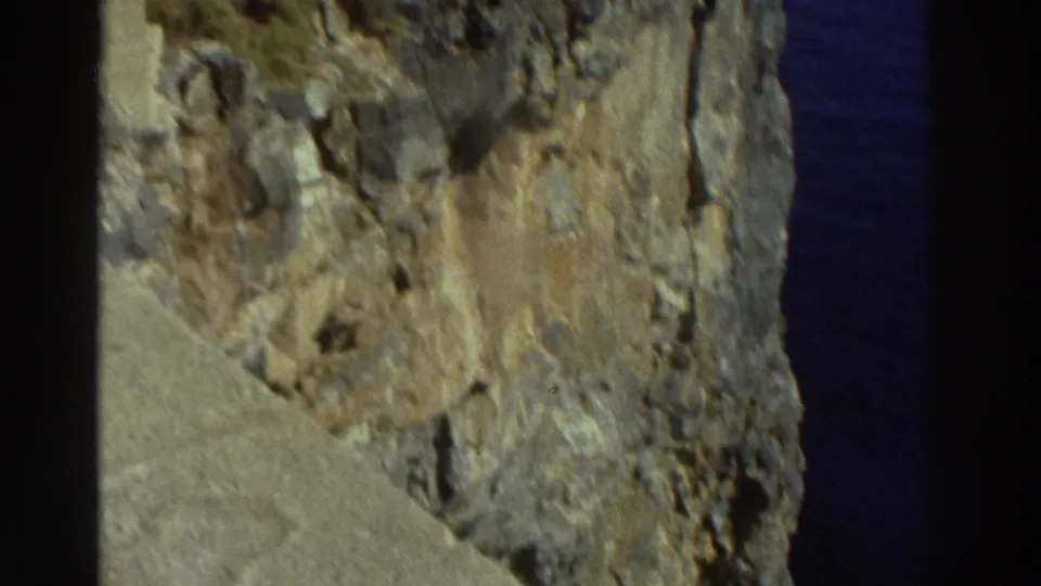 Soft 8mm frame from Greece showing weathered cliff rock beneath stone ruins, with the sea partly visible at right.