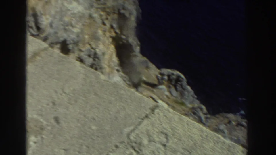 1976 Greece travel footage frame showing a sloped stone surface, rugged cliff rocks, and dark sea below the ruins.