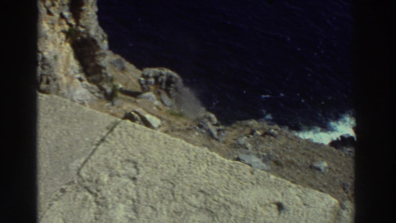 1976 Greece 8mm frame showing rough coastal stone, cliff shadows, dark blue water, and surf breaking against rocks.