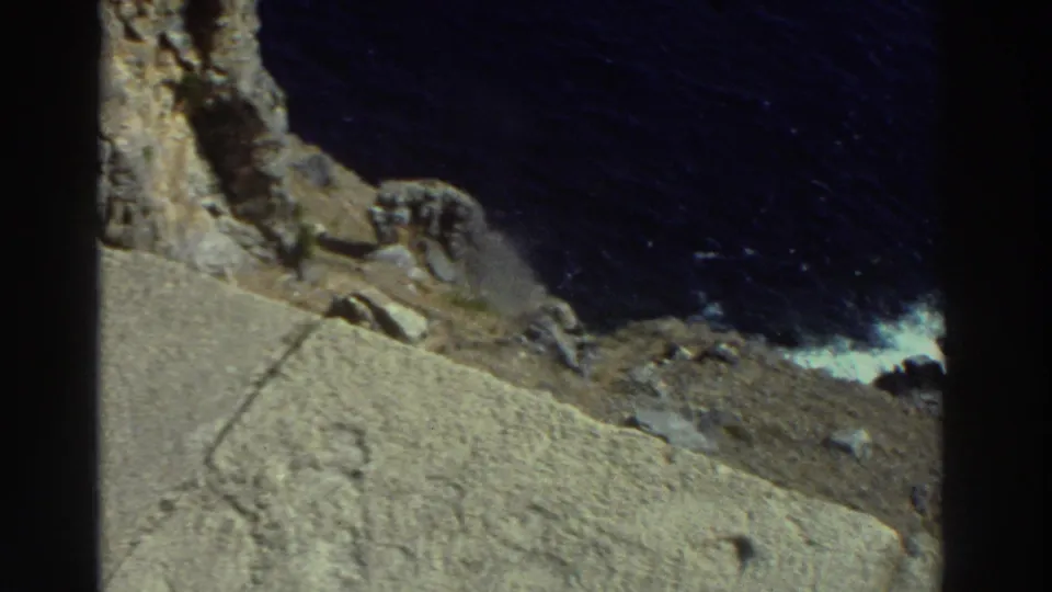 1976 Greece 8mm frame showing rough coastal stone, cliff shadows, dark blue water, and surf breaking against rocks.