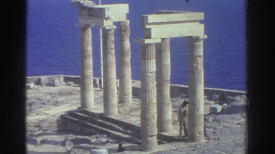 1976 Greece 8mm frame of ancient columns and stepped ruins near the sea, with a distant adult visitor standing beside the structure.
