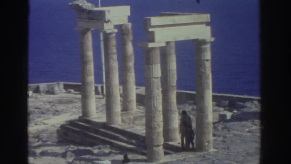 1976 Greece 8mm frame showing ancient columns, a stepped stone base, blue water, and a distant adult visitor in the ruins.