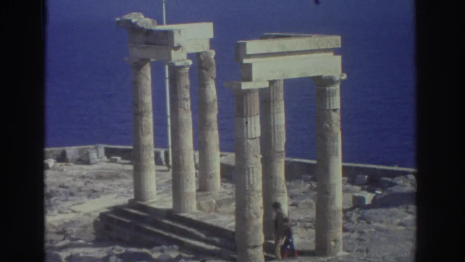 1976 Greece 8mm frame showing seaside ancient columns, stone bases, blue water, and a small visitor figure near the ruins.