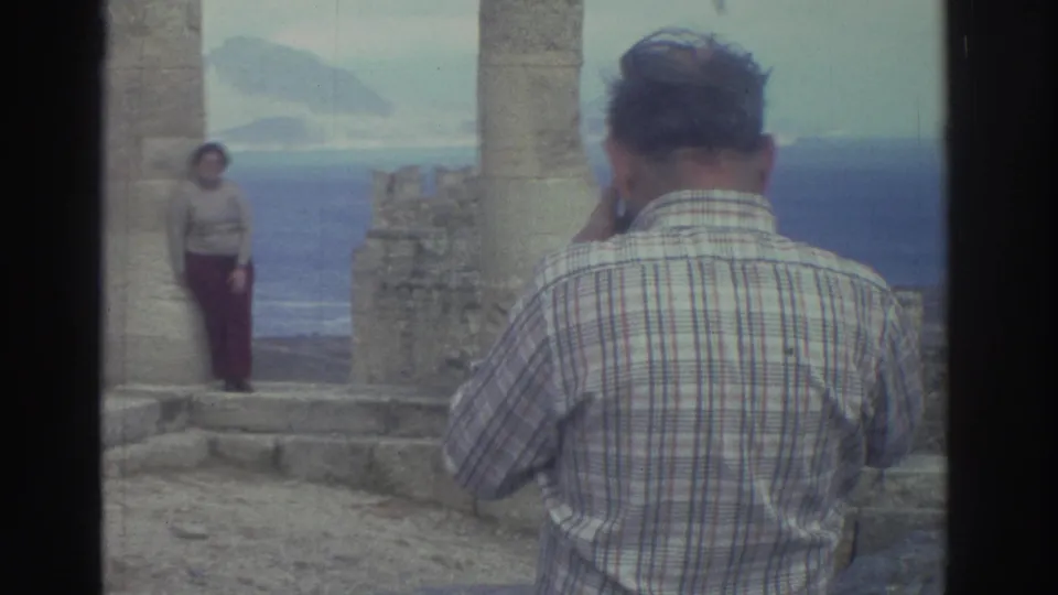 In 1976 Greece, a man in a plaid shirt photographs a woman standing between ancient columns above the sea.