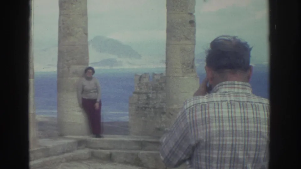 A home-movie view of a tourist photo being taken at Greek ruins, with the woman framed by columns and water behind her.