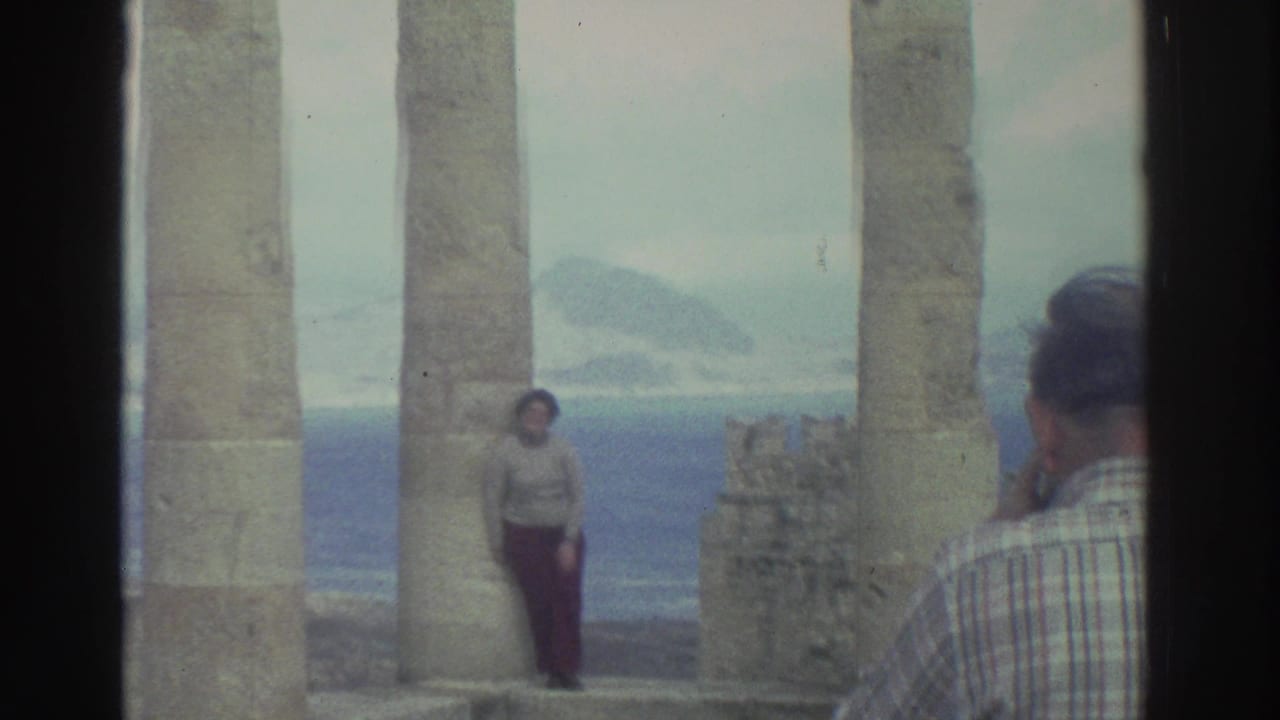 In metadata-supported 1976 Greece, a woman poses among weathered seaside columns with hazy water and landforms behind her.