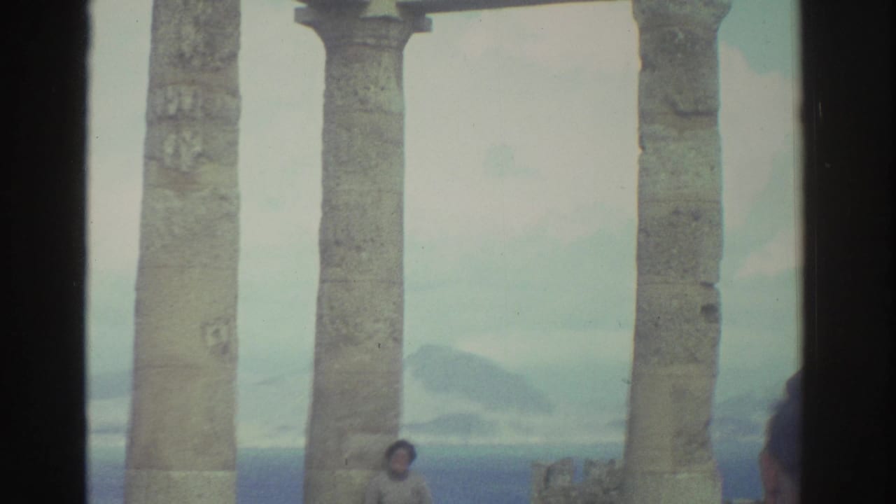 Man in a plaid shirt photographs a woman beside tall stone columns overlooking water.