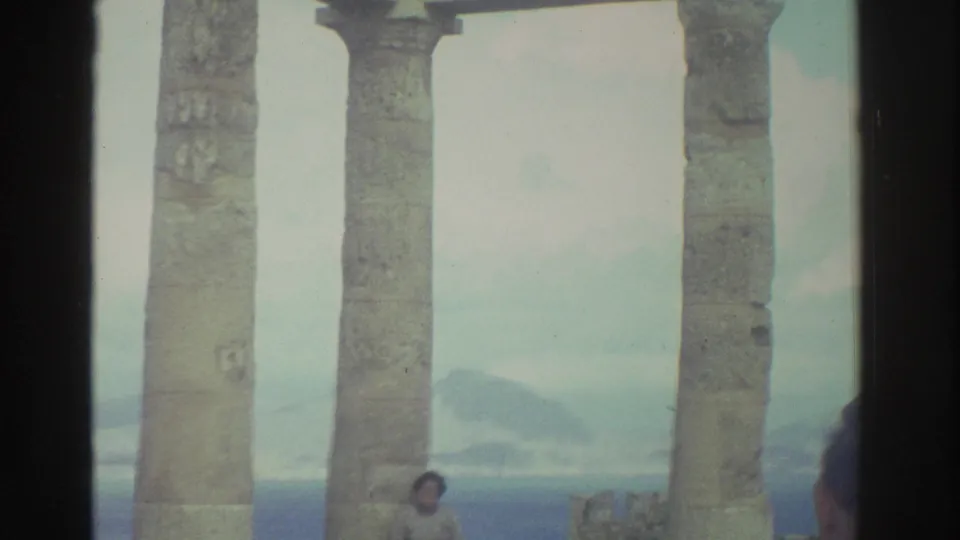 A man in a plaid shirt photographs a woman posing among ancient seaside columns in Greece, with hazy coastal scenery beyond.