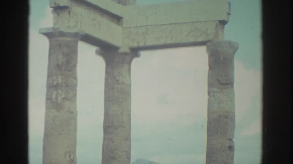 The camera lingers on the weathered column ruins, showing worn stone surfaces and a partial overhead block against the coastal sky.