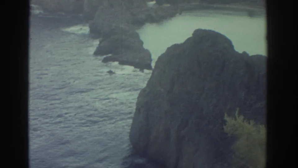 A handheld view shifts from the ruins toward the rocky Greek coastline, showing cliffs, water, and a partly visible visitor at frame edge.