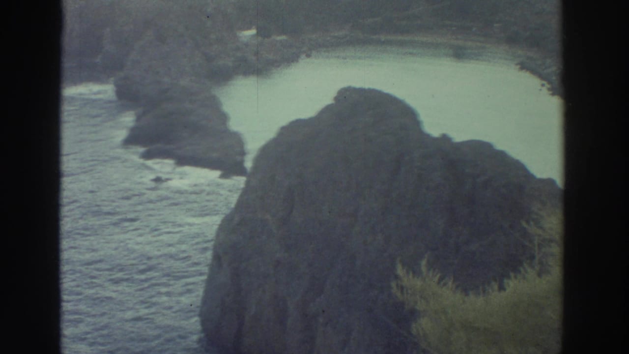 1976 Greece 8mm view of a pale coastal lagoon beside dark rocky formations and sparse shoreline vegetation.