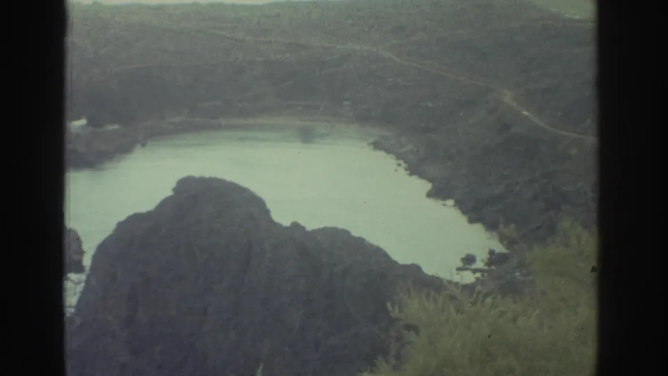 1976 Greece 8mm frame of the lagoon from above, with dark rocks, sparse greenery, and a faint cliffside path.