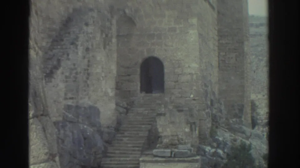 1976 Greece 8mm frame showing a dark arched doorway, stone staircase, high masonry wall, rocky slope, and a small platform.