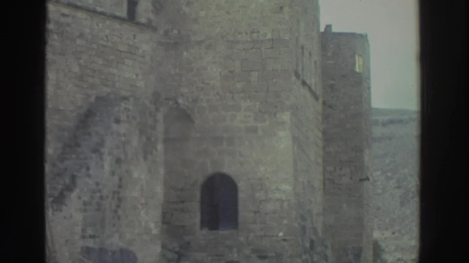 1976 Greece 8mm frame showing castle masonry, a dark arched doorway, a partial tower-like wall, pale sky, and distant hillside.