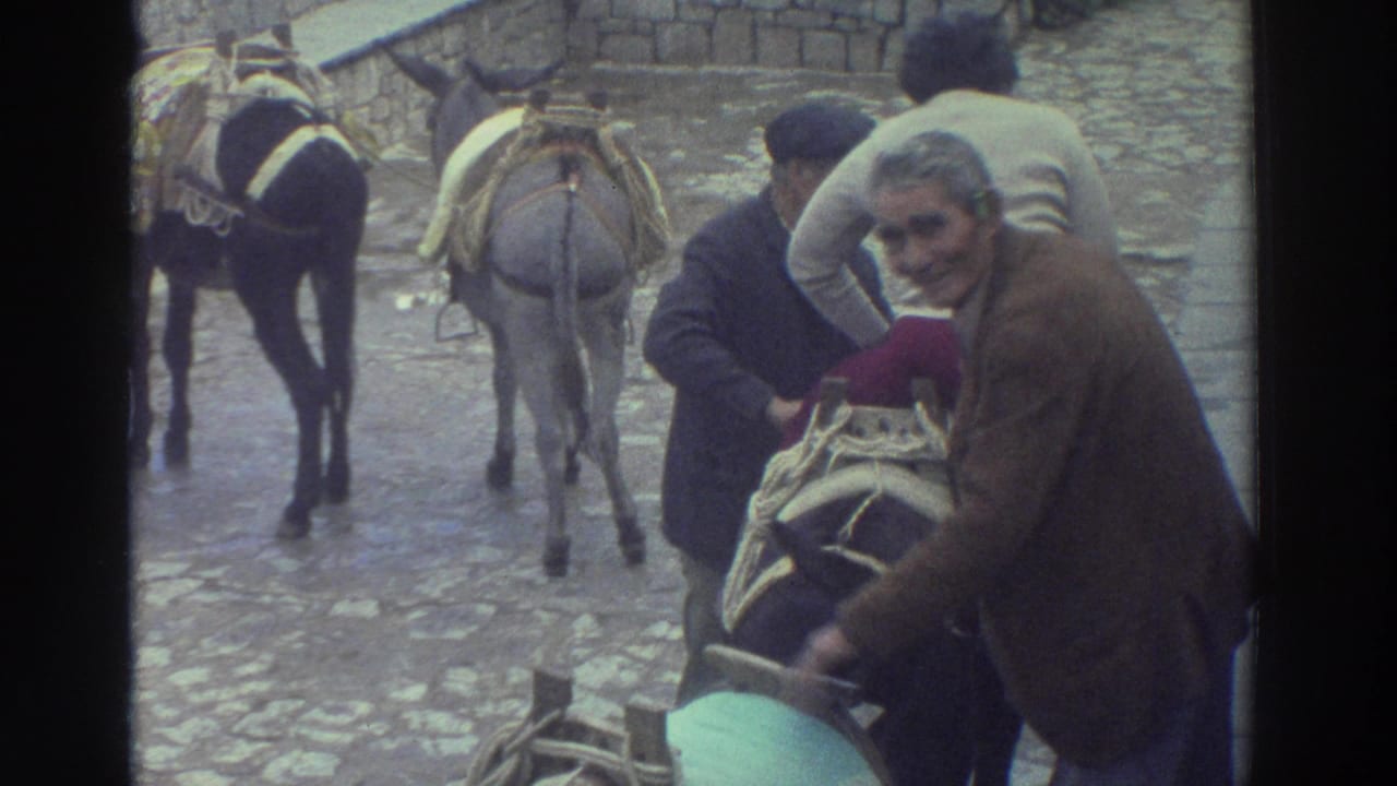 1976 Greece home-movie frame showing adults helping a rider mount a saddled donkey or burro on a stone street.