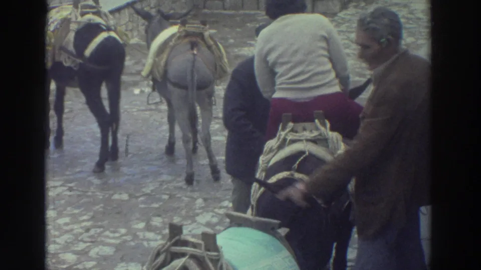 1976 Greece travel footage frame with a seated rider and men adjusting or steadying a saddled donkey or burro.