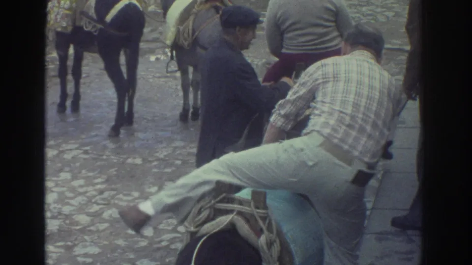 1976 Greece home-movie frame showing a man lifting his leg to mount a saddled donkey or burro on a cobblestone street.