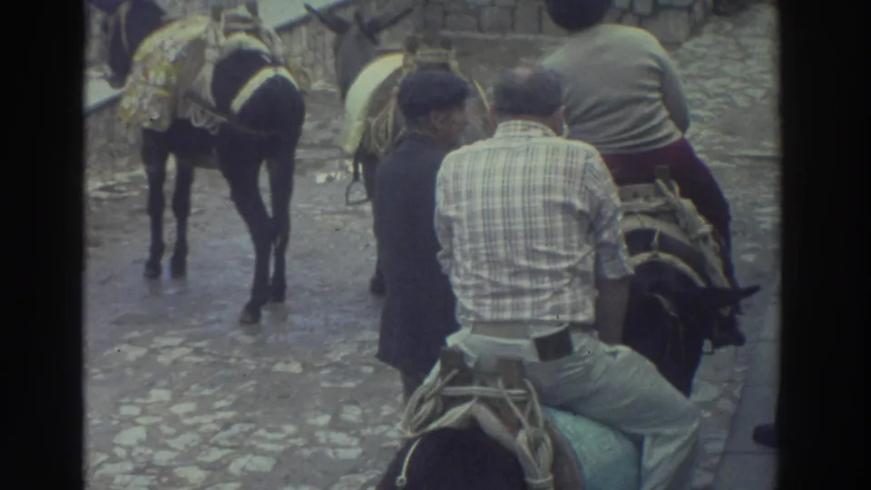 1976 Greece travel frame showing a seated adult on a saddled donkey or burro, with helpers and other packed animals on a stone street.