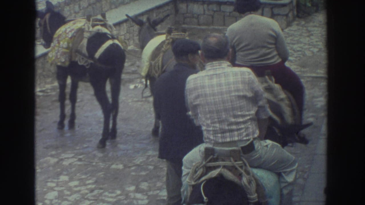 1976 Greece home-movie frame showing men on saddled donkeys moving along a cobblestone path beside a walking guide and low stone wall.