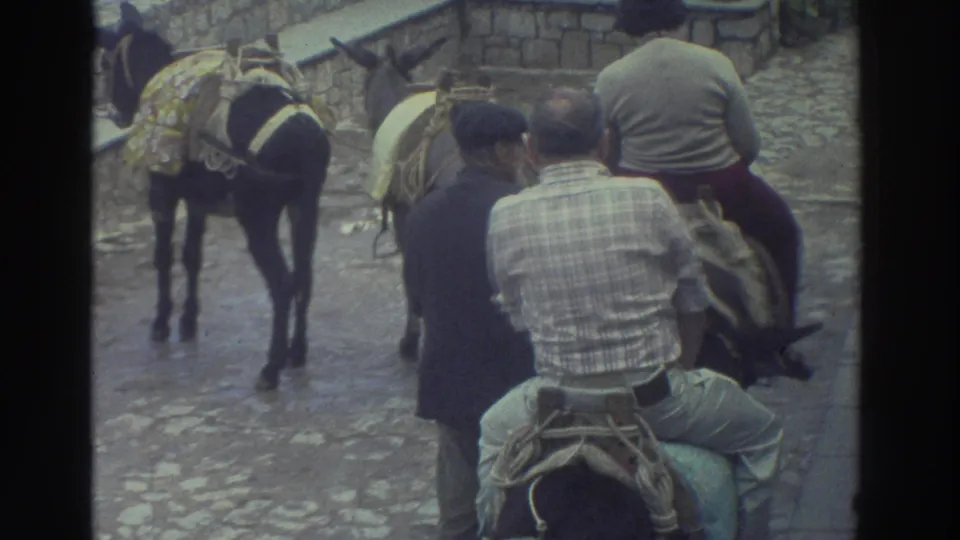1976 Greece home-movie frame showing men on saddled donkeys moving along a cobblestone path beside a walking guide and low stone wall.