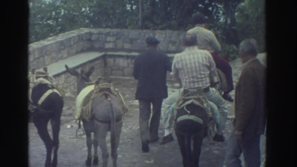1976 Greece home-movie frame showing men on donkeys, a guide walking between them, and an older bystander near a cobblestone path.