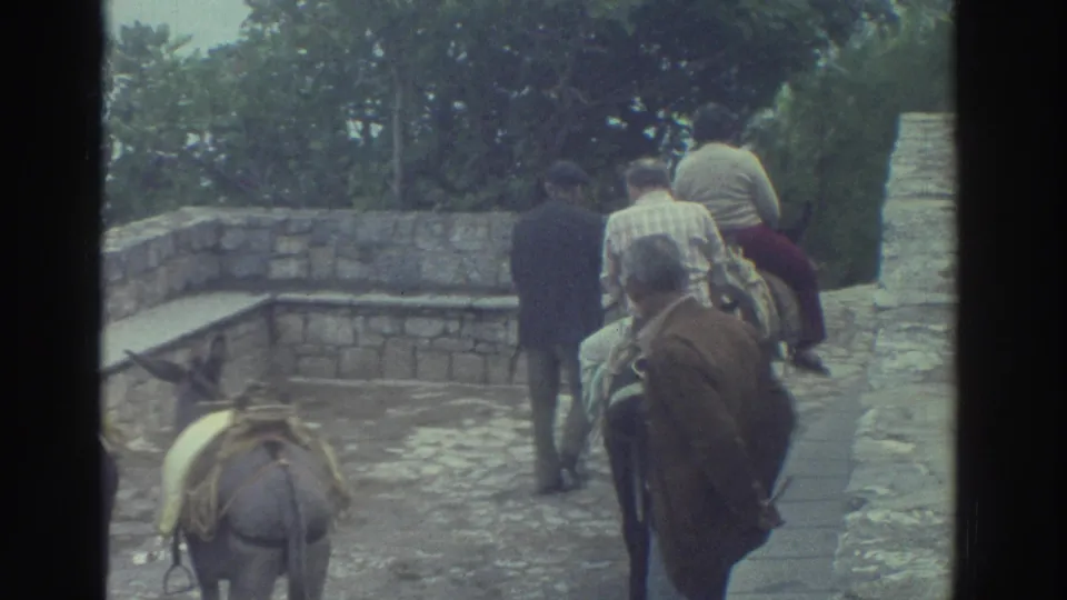 1976 Greece frame showing donkey riders moving away from the camera beside a low stone wall as an older bystander stands in profile.