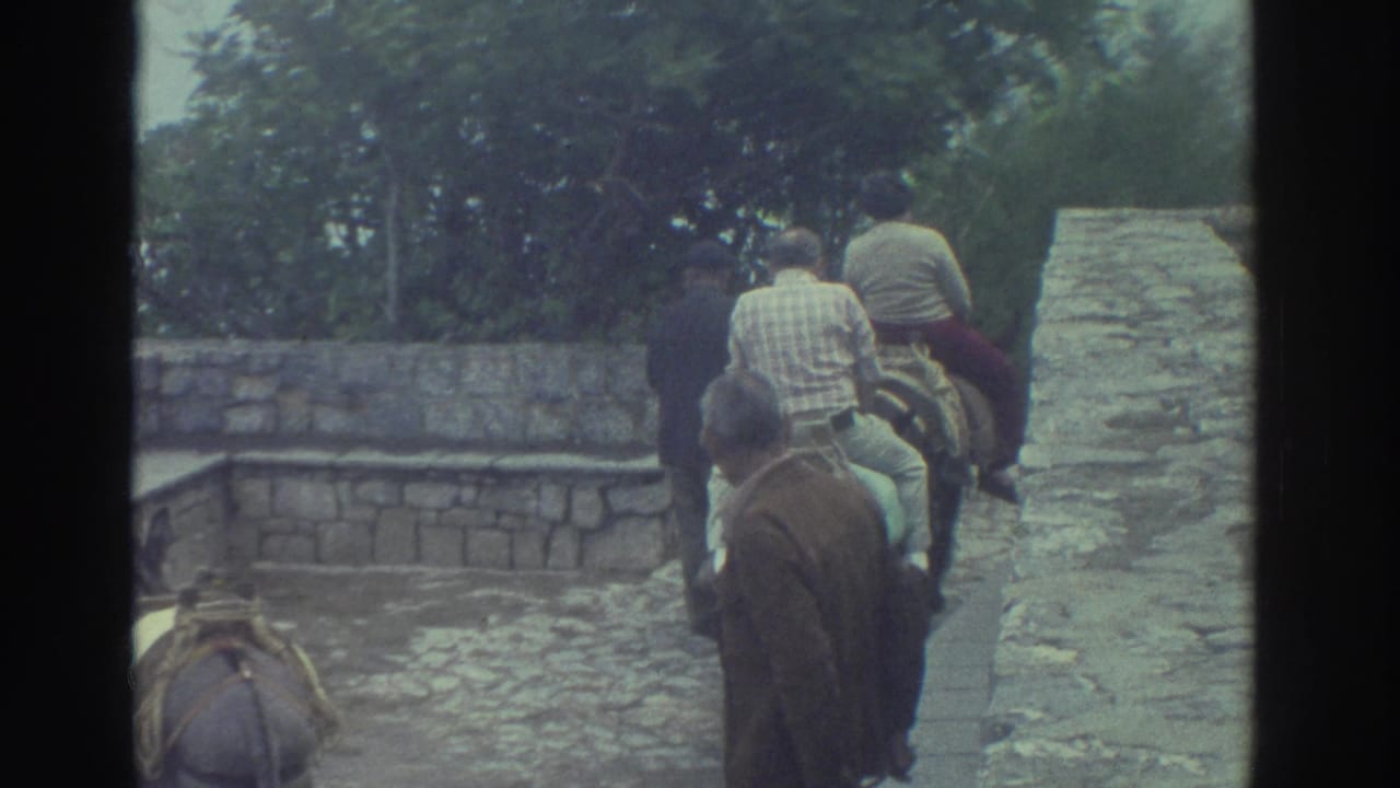 1976 Greece home-movie frame showing people on horseback moving through a stone-paved passage with old masonry walls.