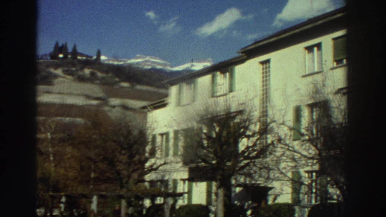 Pale residential building with shutters, bare trees, hills, and snowy mountains in the distance.