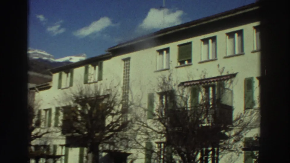 1976 Greece home-movie frame of pale residential facades with green shutters, bare trees, blue sky, and distant mountains.