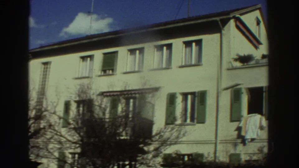 1976 Greece home-movie frame showing a pale residential building with shutters, leafless trees, and hanging laundry near the right edge.