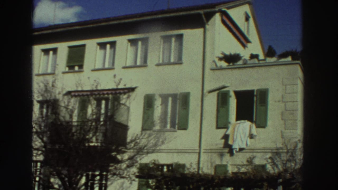 1976 Greece 8mm frame of a white residential building with green shutters, bare trees, and laundry hanging outside a window.