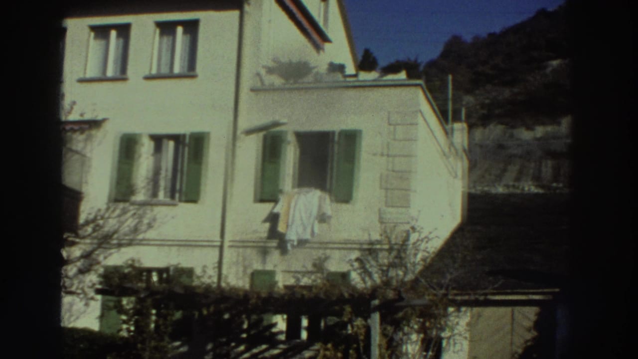 White house facade with green shutters, window laundry, trees, and garden structures.