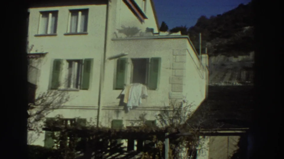 1976 Greece 8mm frame showing the same white residence, green shutters, a hanging cloth, and a shaded garden area below.