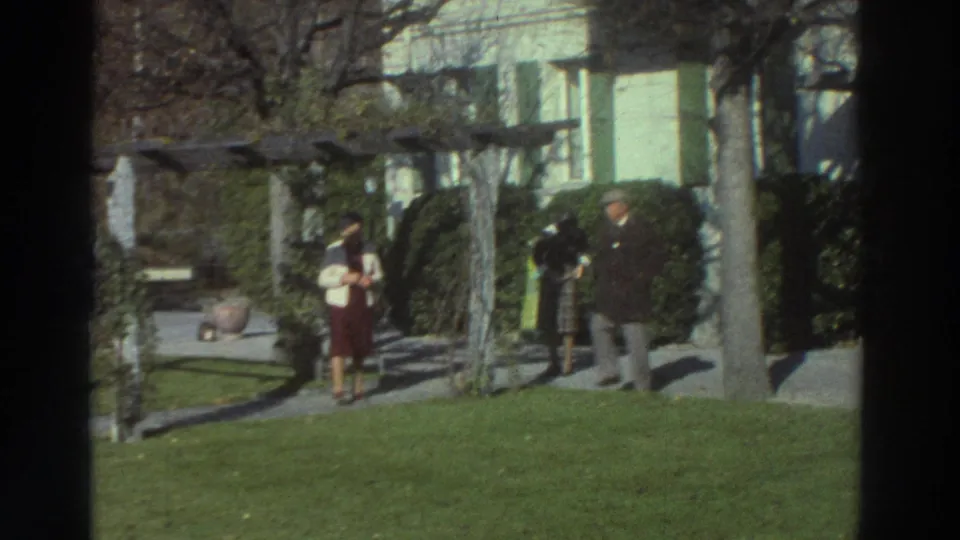 1976 Greece 8mm frame of two adults walking in a sunlit garden, with a pergola, lawn, trees, and house behind them.