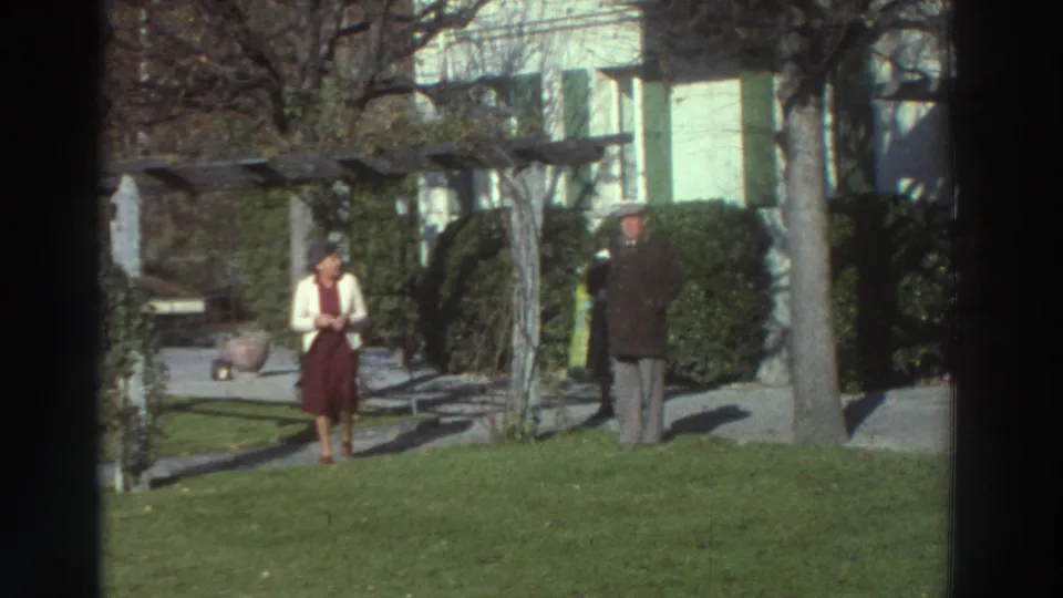 1976 Greece 8mm frame showing a woman in a red dress and a man in a dark coat near shrubs, trees, and a residential garden.