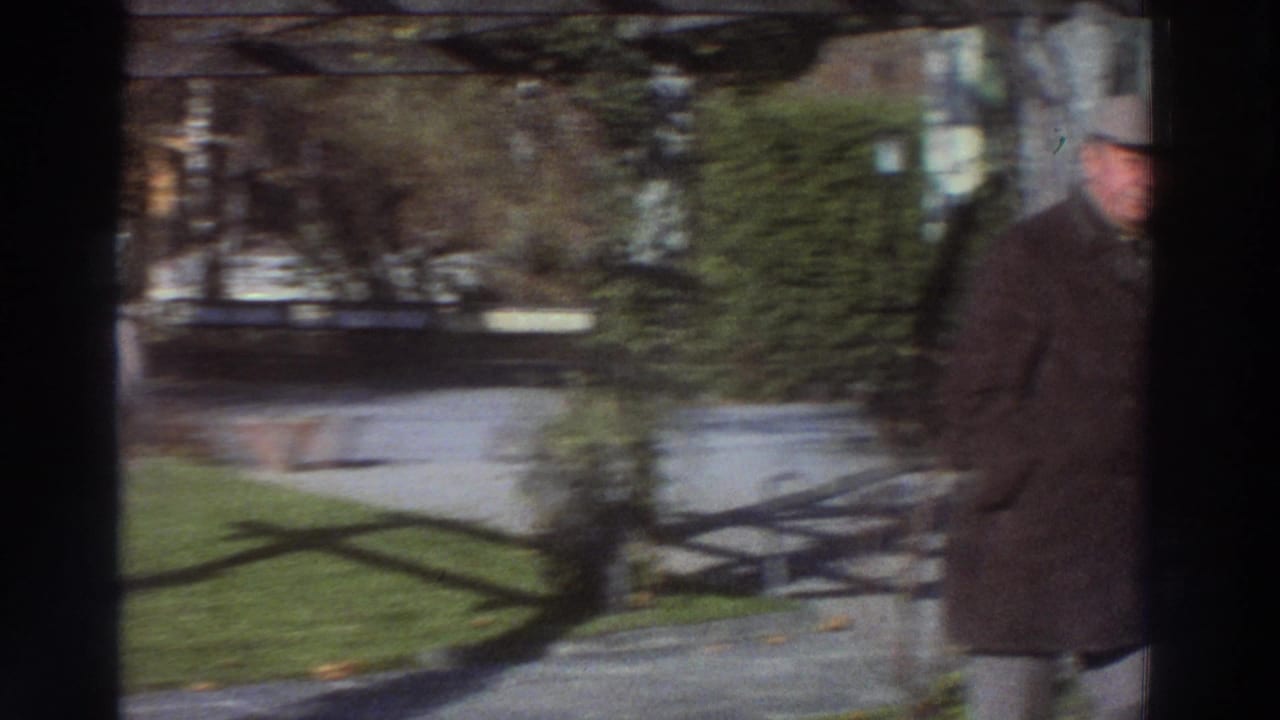 In metadata-supported 1976 Greece, two older men in coats approach along a residential garden path as a woman smiles nearby.