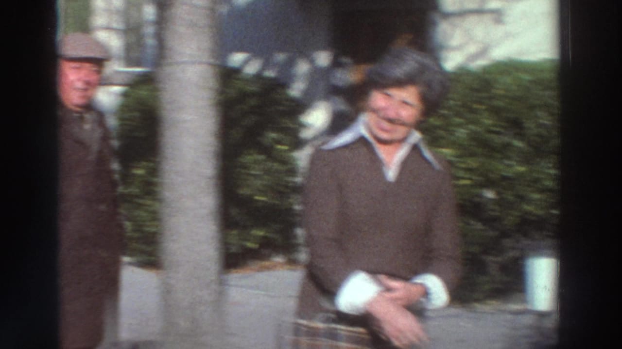 A smiling woman stands beside hedges and a house wall in a garden setting.