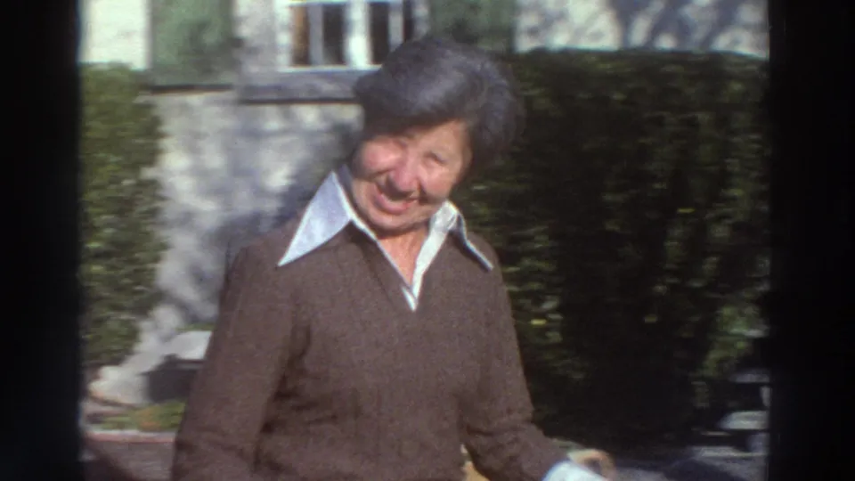 In the 1976 Greece home-movie footage, a woman laughs toward the camera near hedges and a pale house wall.