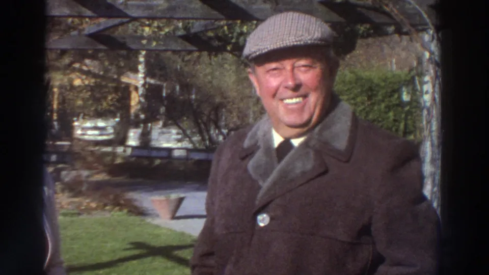 An older man in a brown coat and flat cap smiles toward the camera in the metadata-supported Greece garden setting.