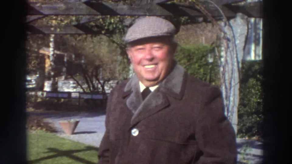 A smiling older man in a cap and dark coat stands outdoors in Greece, based on supplied 1976 location metadata.