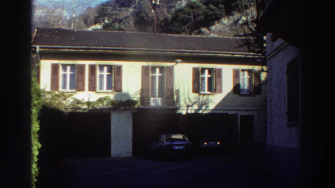1976 Greece home-movie frame showing a shuttered two-story residence with garage openings and classic cars parked outside.