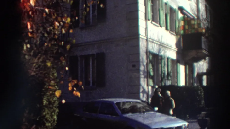1976 Greece home-movie frame showing a multistory residence with green shutters, a balcony, a parked car, and two people near the entrance.