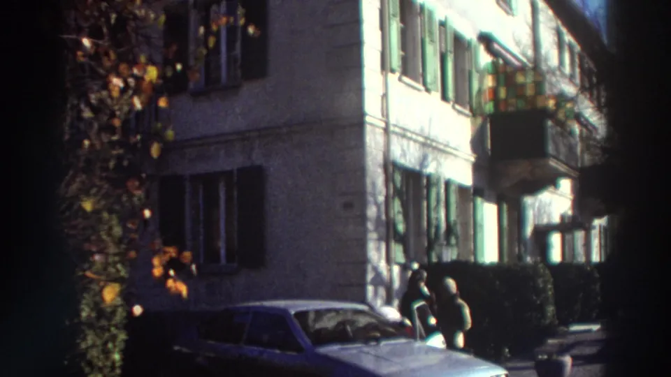 1976 Greece residential street frame with green shutters, a projecting balcony, a classic car, and two people standing near the building.