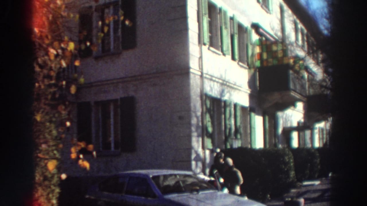 Sunlit residential building with green shutters, parked car, and two figures near the sidewalk.
