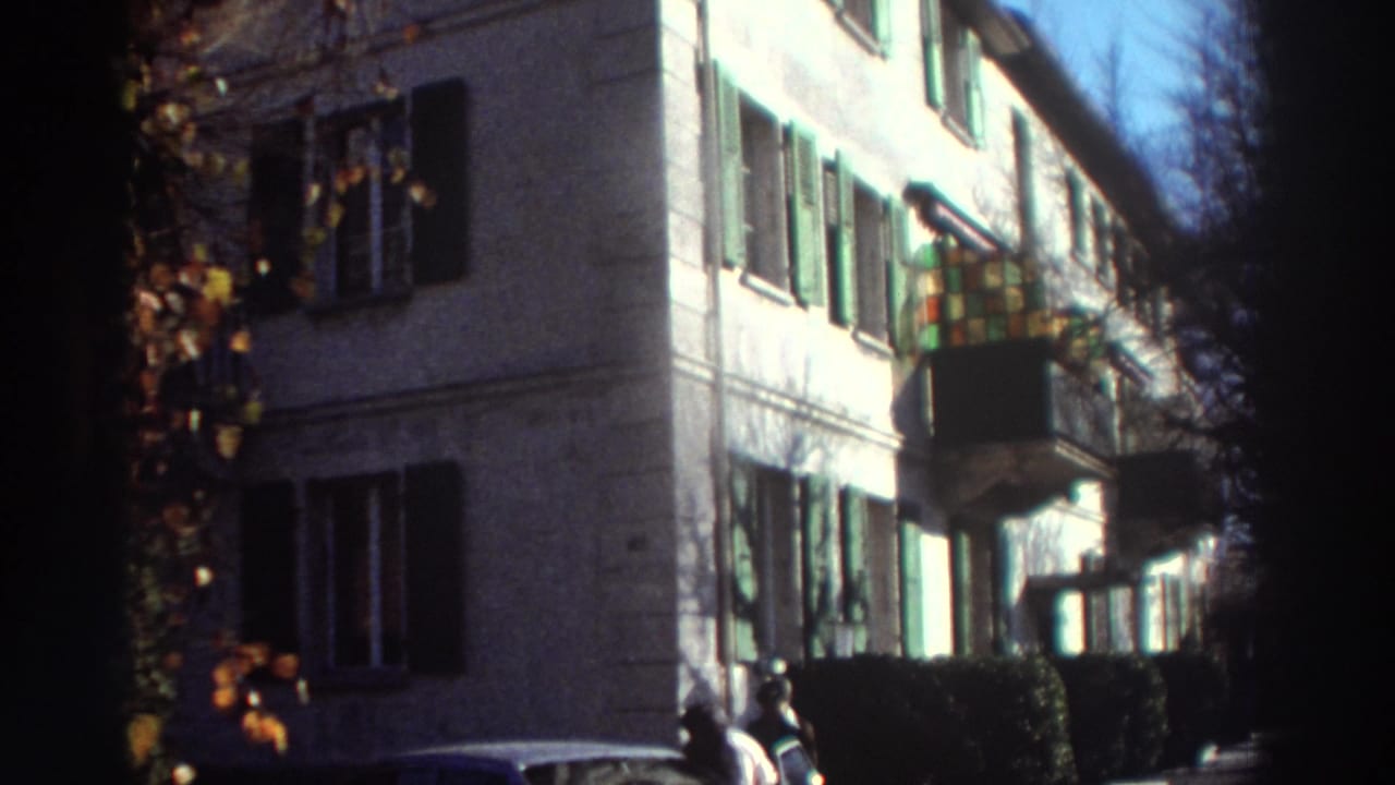 1976 Greece home-movie frame showing a sunlit building facade with green shutters, a small balcony, shrubs, and two people near the street.